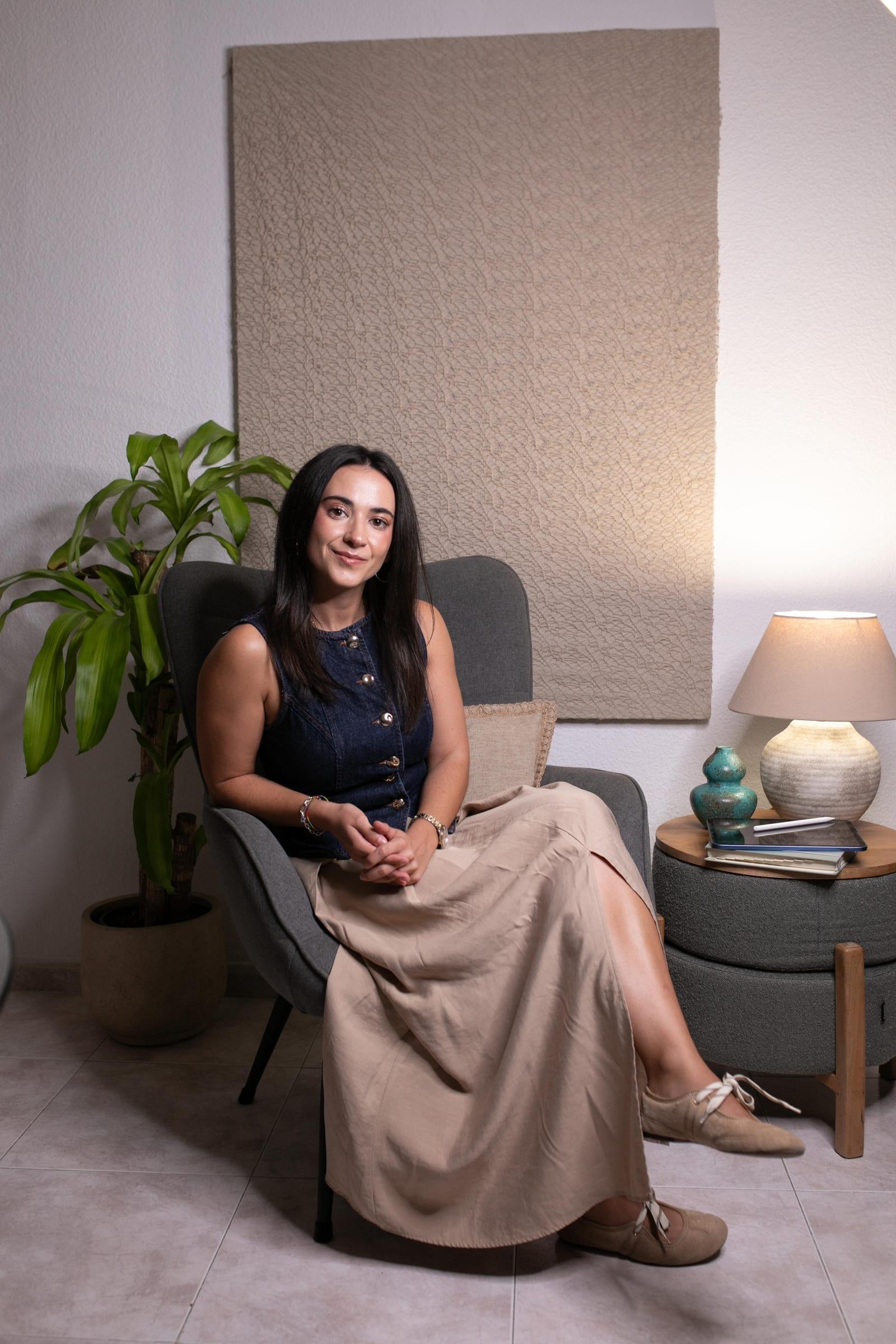 Elena Sánchez Carrasco sentada en un sillón gris de su consulta, junto a una planta y una lámpara encendida